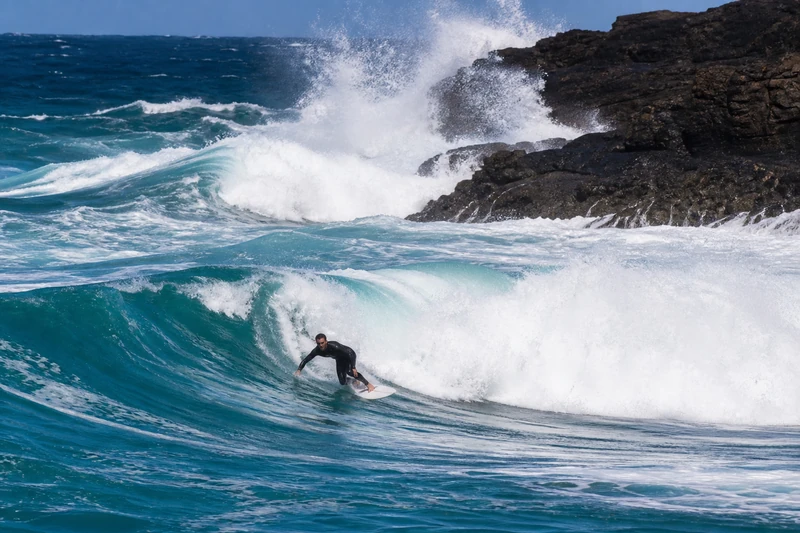 Surfer in a black wetsuit riding a turquoise wave on a white board at Ribeira d'Ilhas, Ericeira, Portugal, with waves crashing against dark cliffs behind