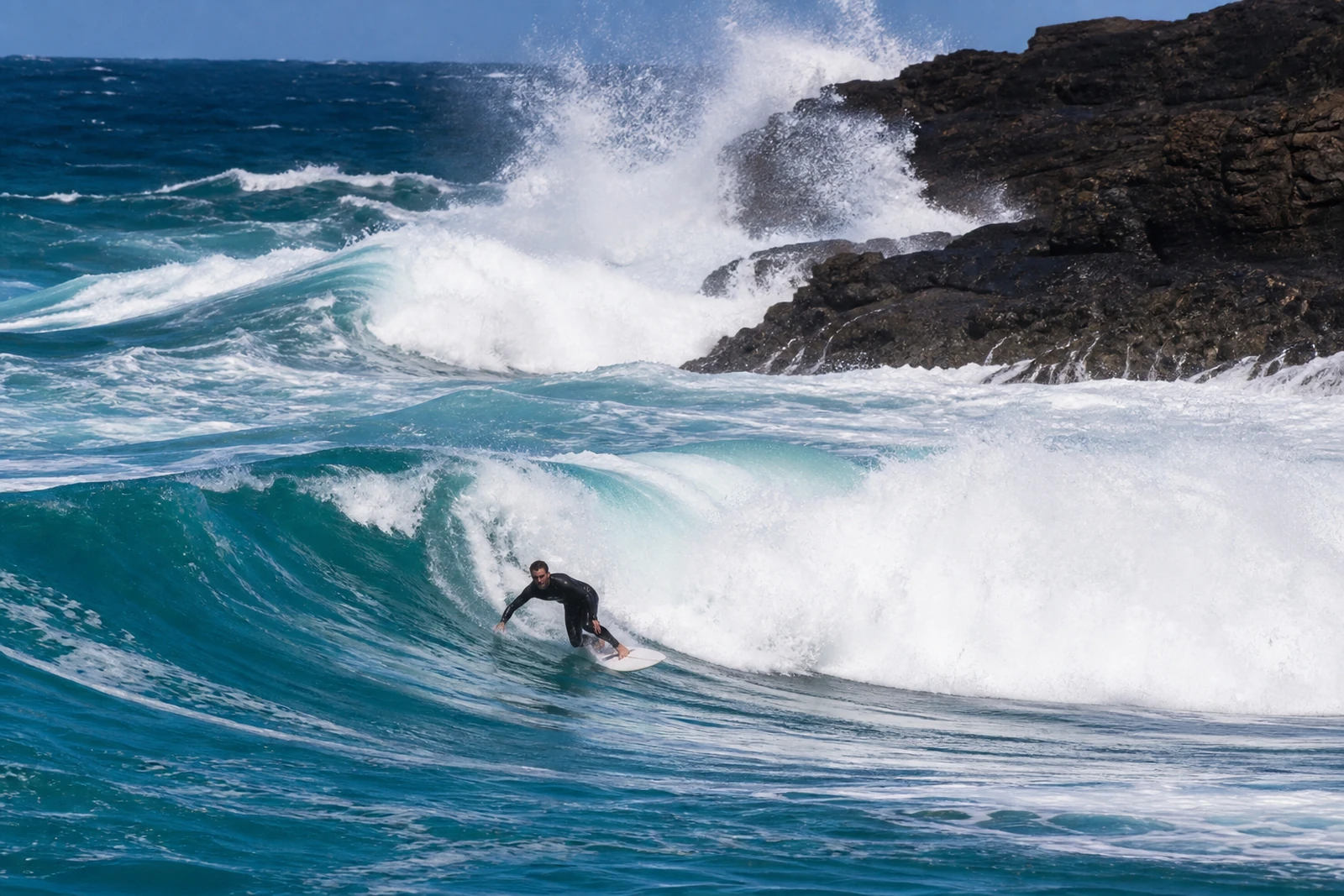 Surfer in a black wetsuit riding a turquoise wave on a white board at Ribeira d'Ilhas, Ericeira, Portugal, with waves crashing against dark cliffs behind